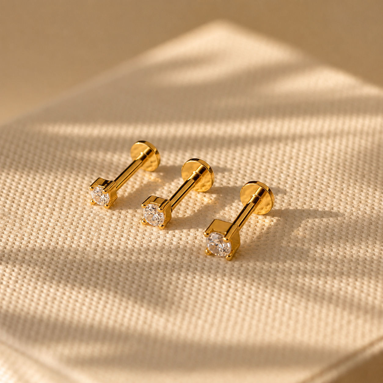 Three gold bar-shaped earrings with clear gemstones on a beige fabric background